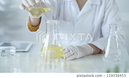 A scientist, wearing a lab coat and white protective gloves, is pouring a yellow oily liquid from one beaker to another in laboratory, close up. Medicine and science concept 119338453