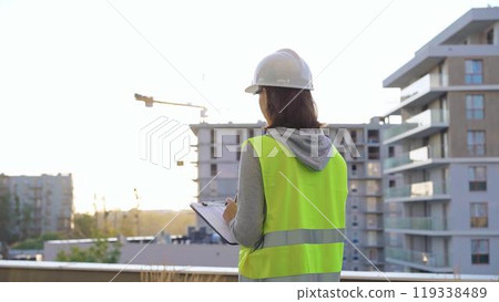 Female engineer with a white protective helmet and safety vest is writing on a clipboard while inspecting a construction site at sunset. Architect concept 119338489