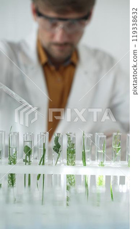 Man scientist wearing a lab coat, white gloves and protective glasses, is dripping liquid from a pipette into a test tube with a green plant inside, vertical portrait. Science and medicine concepts Man scientist wearing a lab coat, white gloves and protective glasses, is dripping liquid from a pipette into a test tube with a green plant inside, vertical portrait. Science and medicine concepts 119338632