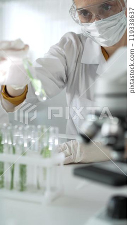 Woman scientist wearing lab coat, white gloves, face mask and protective glasses, is holding a test tube with plants inside near microscope in laboratory, vertical portrait view. Science and medicine 119338637