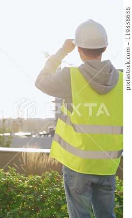 Man engineer with white helmet and safety vest is using tablet computer while inspecting a construction site at sunrise, back vertical view 119338638