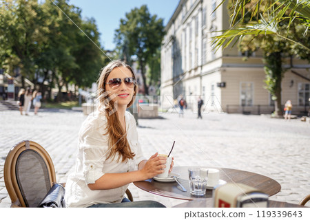 Young woman enjoying coffee at a cafe in a vibrant outdoor square on a sunny day 119339643