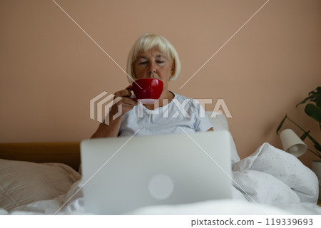 Female using laptop. Caucasian senior 60s 70s woman sitting with cup of coffee on bed and having breakfast while enjoying morning in bedroom at home. Morning rituals. 119339693