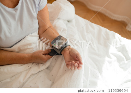 Close up of woman checks her blood pressure, a tonometer is worn on her wrist in a bed. Medical device for measuring blood pressure and heart rate used at hand wrist 119339694