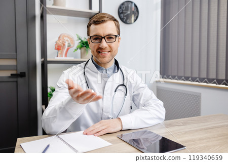 Portrait of young doctor wearing glasses and white uniform with stethoscope, speaking and consulting patient online, looking at camera, making video call, sitting at table in office Portrait of young doctor wearing glasses and white uniform with stethoscope, speaking and consulting patient online, looking at camera, making video call, sitting at table in office 119340659