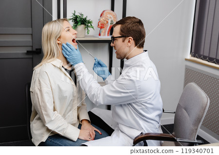 Doctor using inspection spatula to examine patient throat. ENT doctor doing throat exam of a woman. Patient opened her mouth to throat check-up 119340701