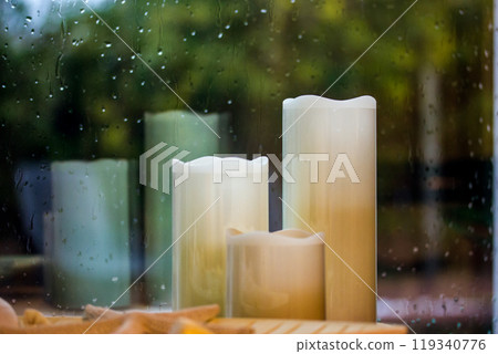 Set of white yellow paraffin artificial tall candles against wet window with water droplets, fall rain outside a window Decor for cozy home comfort. Autumnal cold rainy weather still life. Fall season 119340776