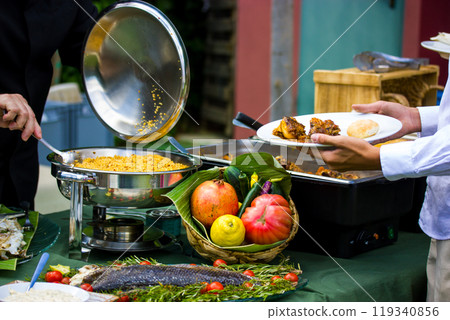 A chef cook puts freshly cooked food on a plate. Stainless steel buffet container, Food tray. Tables set at a banquet. A fruit and vegetable composition. Fresh fruits, vegetables, fish, salat couscous 119340856