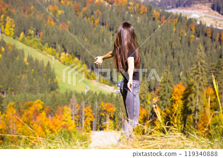 Free happy young woman with long brown hair in jeans and a T-shirt stands on top of a mountain with arms outstretched to her sides. A traveler, a tourist, an adventurer. Hiking female in Ukraine. Free happy young woman with long brown hair in jeans and a T-shirt stands on top of a mountain with arms outstretched to her sides. A traveler, a tourist, an adventurer. Hiking female in Ukraine. 119340888
