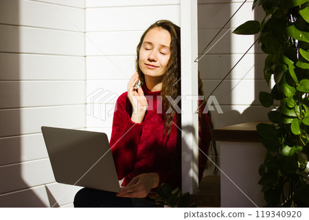 Happy calm beautiful girl smiling, exposed face to the sun working at laptop in a comfort home. Young woman works remotely in cozy workplace indoors in a house, cottage. Positive vibes real emotions. 119340920