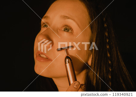 Young beautiful white woman, girl is using vibrating golden facial massager on dark black background with copy space. Preservation prolongation of natural beauty youth Female skin care with pleasure. Young beautiful white woman, girl is using vibrating golden facial massager on dark black background with copy space. Preservation prolongation of natural beauty youth Female skin care with pleasure. 119340921