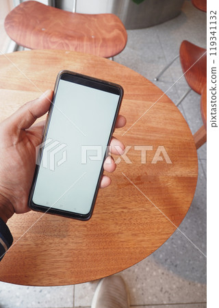 A hand is casually holding a smartphone while resting on a wooden tables surface A hand is casually holding a smartphone while resting on a wooden tables surface 119341132
