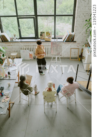 Vertical high angle shot of teacher giving drawing lesson to senior people in spacious studio with huge rectangular windows Vertical high angle shot of teacher giving drawing lesson to senior people in spacious studio with huge rectangular windows 119341223