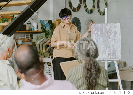 Back view shot of senior biracial group of people listening to young female teacher who explaining drawing tips in front of student group in studio Back view shot of senior biracial group of people listening to young female teacher who explaining drawing tips in front of student group in studio 119341300
