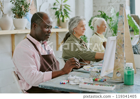 Small group of biracial retired people painting scenery on canvases during art therapy together 119341405