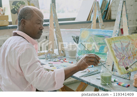 Senior African American man sitting at dirty desk with painting tools and wetting paintbrush in jar with dirty water 119341409