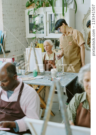 Vertical shot of young adult drawing teacher checking her senior student who trying to study new drawing technique in background Vertical shot of young adult drawing teacher checking her senior student who trying to study new drawing technique in background 119341507