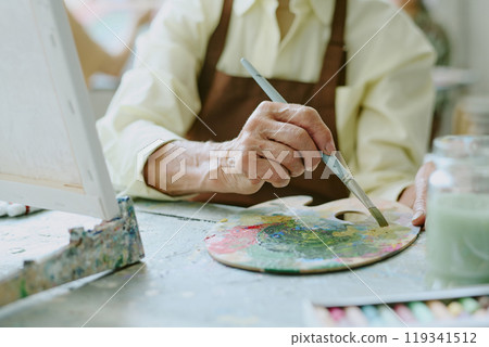 Medium close up of unrecognizable senior female woman holding brush while mixing oil paints on dirty wooden palette 119341512