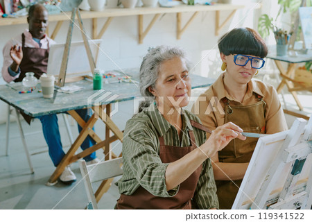 High angle shot of group of senior people drawing with oils paints on canvas while their young female teacher watching them High angle shot of group of senior people drawing with oils paints on canvas while their young female teacher watching them 119341522