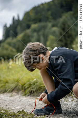 Young boy kneeling on a footpath in green nature to tie his hiking shoes 119341543