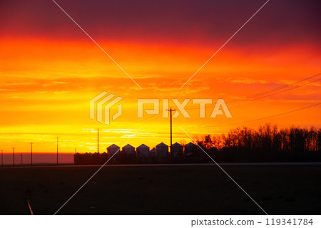 Bright beautiful sunrise with orange and purple light along the highway and silhouettes of a grain bins at the farm in the horizon in autumn. Bright beautiful sunrise with orange and purple light along the highway and silhouettes of a grain bins at the farm in the horizon in autumn. 119341784