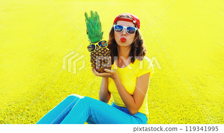 Summer portrait happy young woman blowing kiss with pineapple, glasses on the grass in summer park 119341995