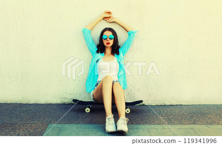 Portrait of beautiful young woman with skateboard wearing blue shirt, sunglasses on street 119341996