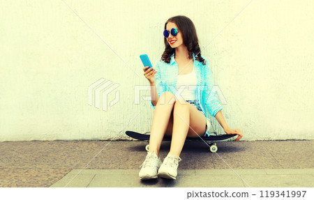 Modern young woman with smartphone sitting on skateboard on city street on white background 119341997
