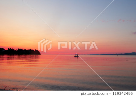 Idyllic morning at the ocean shore, calm waters, orange and pink sunrise and a silhouette of a  man on a paddle boat, mountains in the background. 119342496