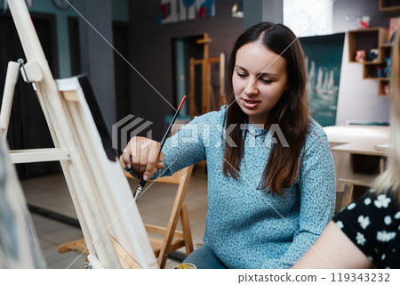 Young woman painting on canvas in a bright art studio during daytime Young woman painting on canvas in a bright art studio during daytime 119343232