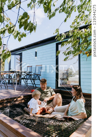 Family enjoying outdoor relaxation on a sunny day, playing on a net hammock outside a modern house Family enjoying outdoor relaxation on a sunny day, playing on a net hammock outside a modern house 119344275