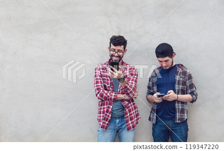 Two smiling friends leaning on a wall checking their cell phones, Friends leaning on a wall texting on their phones. Friend showing cell phone to his friend, Smiling friends checking cell phones 119347220