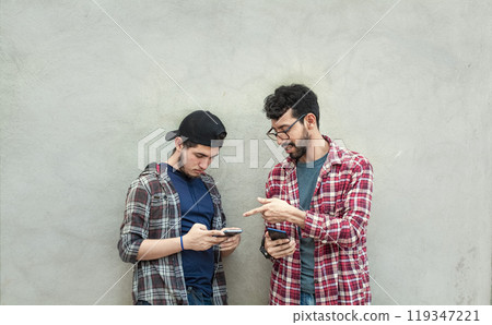 Two young friends leaning against a wall checking their cell phones, Two friends leaning against a wall looking at the content on their phones. Friend showing cell phone to his friend outdoors 119347221