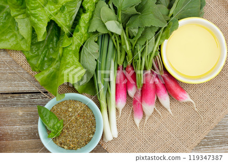 Fresh ripe vegetables on the wooden table with burlap table cloth - radish, green onion, lettuce, dried herbs in a little prep bowl with basil leaves and olive oil are ready to cook salad. 119347387