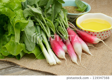 Fresh ripe vegetables on the wooden table with burlap table cloth - radish, green onion, lettuce, dried herbs in a little prep bowl with basil leaves and olive oil are ready to cook salad. 119347388