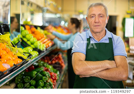 Portrait of confident mature male supermarket worker wearing apron standing in middle of grocery store 119349297