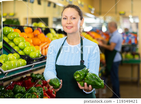 Professional adult female seller in apron putting red and green bell peppers on stalls in hypermarket Professional adult female seller in apron putting red and green bell peppers on stalls in hypermarket 119349421