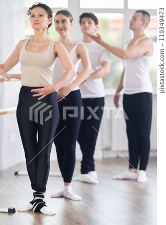 Group of young dancers engaged in ballet in dance studio perfoms exercise near ballet barre, male choreographer helps to coordinate movements correctly 119349473
