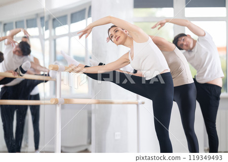 Beautiful athletic girl with group of dancers practicing ballet pose grand batman near ballet barre in dancing hall 119349493