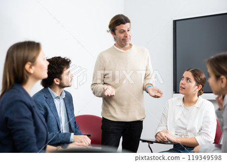Middle-aged man business coach gesturing with hand while standing against group of people sitting in circle in auditory Middle-aged man business coach gesturing with hand while standing against group of people sitting in circle in auditory 119349598