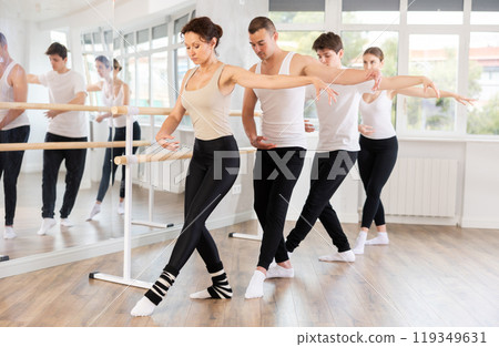 Group of positive women and men standing in row together while doing ballet dance moves dancers standing with one hand on barre and reaching over hand in fitness room 119349631