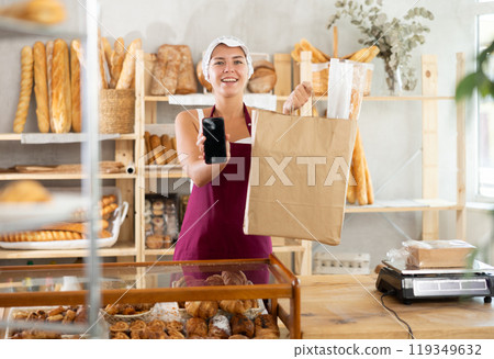 Young saleswoman holds phone and paper bag with baguettes Young saleswoman holds phone and paper bag with baguettes 119349632