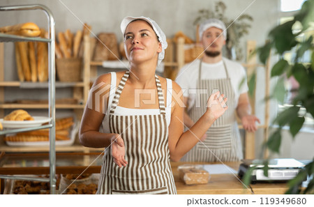 Content young girl worker stands with her back to frustrated man behind bakery counter Content young girl worker stands with her back to frustrated man behind bakery counter 119349680