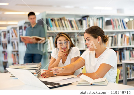 Positive female students using laptop and talking with each other in the book space library 119349715