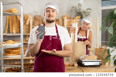Guy seller in bakery hold out hand with phone, points at screen with finger Guy seller in bakery hold out hand with phone, points at screen with finger 119349739