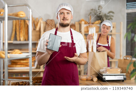 Guy seller in bakery hold out hand with phone, points at screen with finger Guy seller in bakery hold out hand with phone, points at screen with finger 119349740