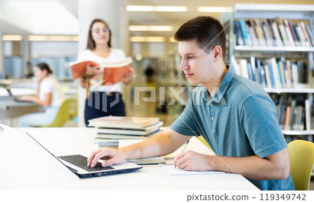Portrait of positive young man with laptop and book in library Portrait of positive young man with laptop and book in library 119349742