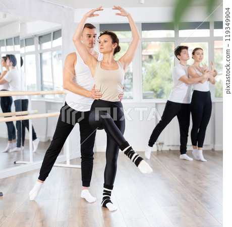 Smiling young man and female partner in black and white clothing with group active people training ballet dance movements in dance salon Smiling young man and female partner in black and white clothing with group active people training ballet dance movements in dance salon 119349904