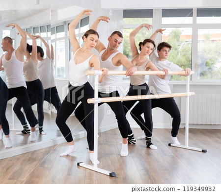 Group of sports people standing in row together while doing ballet dance moves demi plie while holding on ballet barre in ballet studio Group of sports people standing in row together while doing ballet dance moves demi plie while holding on ballet barre in ballet studio 119349932