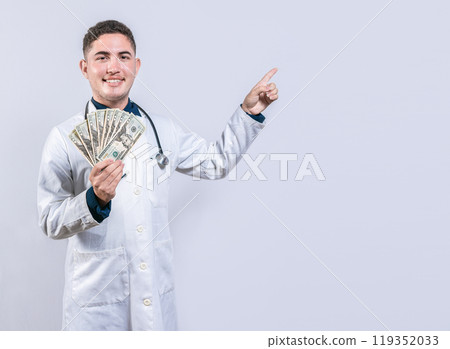 Happy latin doctor holding money and pointing to side. Handsome smiling doctor showing money and pointing side 119352033